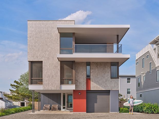 Contemporary multi-storey residential house with a flat roof, projecting upper-level balcony, pale shingle-clad walls, dark grey garage door, and a bold red entry section. The facade combines large glazed openings, metal or glass balcony balustrades, and slender structural supports. Adjacent coastal-style homes and a person carrying a surfboard are visible in the surroundings.