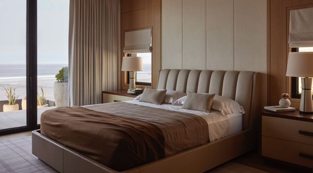 A refined bedroom with a large upholstered bed, padded channel-tufted headboard, warm wood wall panelling, coordinated bedside tables, table lamps, and layered neutral bedding. Full-height glazing and curtains open to a coastal view, while a tiled or stone-look floor continues throughout the room. The scheme is warm, restrained, and luxurious.
