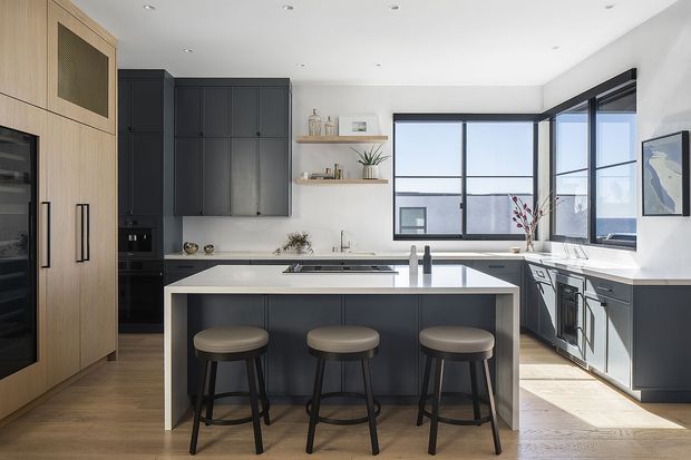 A bright contemporary kitchen with a large central island and three stools. The room features dark charcoal cabinetry, light timber pantry-style joinery, white benchtops and splashback surfaces, integrated appliances, recessed ceiling lights, and expansive black-framed corner windows with an outdoor view.