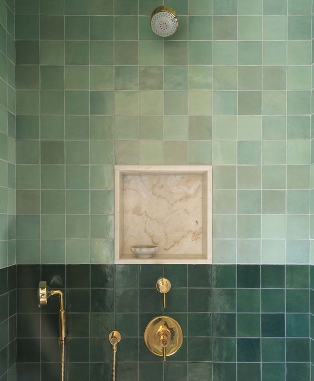 Close-up view of a tiled shower wall with a handheld and overhead shower fitting, brass controls, and a recessed marble-lined niche with a small soap dish. The wall combines pale muted green square tiles above with darker glossy green tiles below, separated by a horizontal transition.