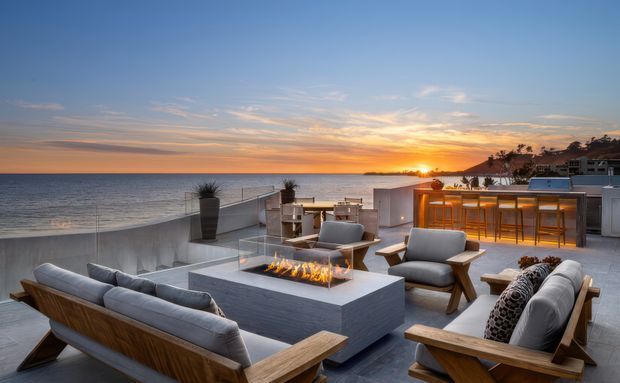 A contemporary rooftop or elevated seaside outdoor lounge overlooking the ocean at sunset. The terrace features timber-framed upholstered lounge seating, a large square concrete fire table with flames, glass balustrades, and a built-in outdoor bar with warm illuminated cabinetry. Modern coastal architecture and a dramatic horizon sunset define the scene.
