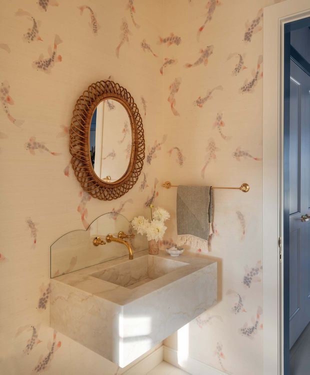 A compact, warmly lit bathroom with a floating rectangular stone vanity and integrated basin, brass wall-mounted tapware, a clear glass splash guard, and a brass towel rail. The walls are covered in cream wallpaper with a delicate botanical and grape-like pattern. An ornate woven rattan-framed oval mirror hangs above the vanity, and a blue-painted door is visible at right.