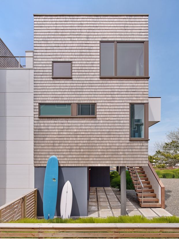 Exterior view of a narrow multi-storey contemporary coastal residence. The main facade is clad in small, horizontally coursed pale timber shingles with dark trim around varied rectangular windows. An elevated upper volume sits above a partially open ground level, with an external timber stair, paved entry courtyard, surfboards, planted grasses, and a slatted timber foreground fence.