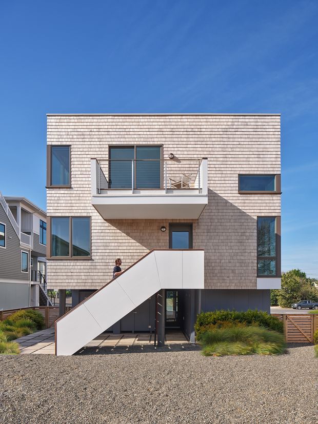 A contemporary multi-storey residential building photographed from the street, featuring a rectangular shingle-clad facade, large dark-framed windows, an upper balcony with metal railing, and a prominent exterior stair rising to the entry. The lower level is dark and recessed, with gravel landscaping, grasses, timber fencing, and neighbouring homes visible around it under a clear blue sky.