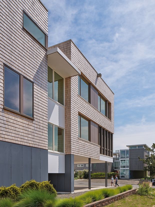 Exterior architectural photograph of a contemporary multi-storey building with a strongly articulated facade clad in small light-coloured shingle-like tiles, dark lower wall panels, large glazed windows, projecting balconies and recessed entry or circulation spaces. A landscaped forecourt and neighbouring modern buildings are visible under a partly cloudy sky.