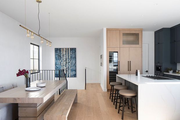 A bright, open-plan dining and kitchen interior with pale timber flooring and a large pale stone island. The dining area has a long timber table with bench seating, while the kitchen combines dark navy cabinetry with a light timber appliance wall and integrated appliances. Black-framed glazing, a black metal stair balustrade, minimalist pendant lighting, and a blue-toned forest artwork add contrast. The space has a clean, refined contemporary feel.