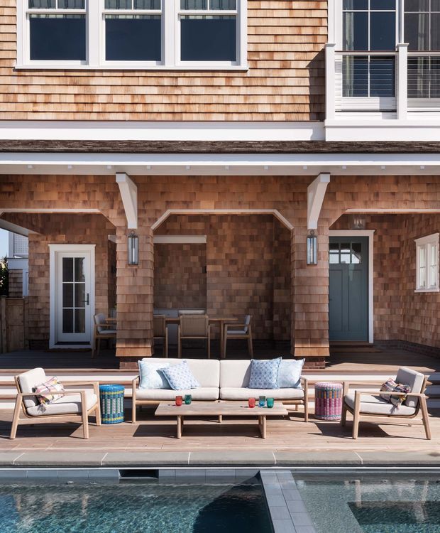 A detailed exterior view of a multi-level shingle-clad home overlooking a swimming pool. The facade features warm timber shingles, white-trimmed windows, a covered ground-level outdoor entertaining area with timber columns, and a dark blue-grey entry door. A timber pool deck is furnished with a light outdoor sofa, lounge chairs, coffee table, and colourful side stools.