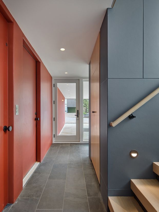 A narrow contemporary hallway and stairway with bold terracotta-red built-in doors and wall panels on the left, dark grey wall panelling on the right, and a light timber handrail and stair treads. Large-format grey floor tiles lead to a glazed exterior door at the end of the corridor. Recessed ceiling lights and a small circular wall light provide understated illumination.