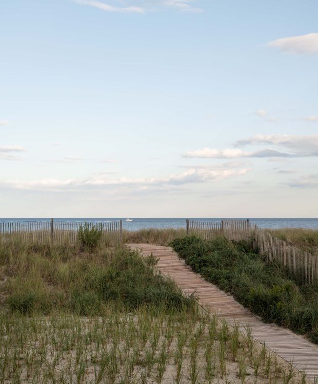 A sandy coastal dune landscape with a narrow timber boardwalk leading through beach grasses toward the ocean. Slender timber fencing lines both sides near the shoreline, with a calm blue sea and expansive pale sky beyond. The scene is natural, open, and serene, with no building visible.