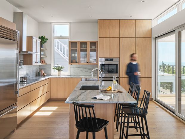 A bright, spacious contemporary kitchen with extensive warm timber cabinetry, a large marble-look island with integrated sink and seating, stainless-steel appliances, and black spindle-back bar stools. Full-height sliding glass doors open to an exterior terrace or balcony, while a stairwell and glazed display cabinetry are visible beyond the main kitchen. Natural light, pale timber flooring, and streamlined recessed lighting create an airy, refined interior.