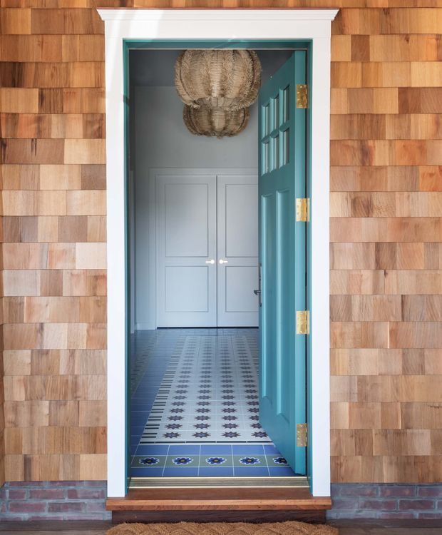 A welcoming exterior entryway viewed straight on, framed by warm cedar shingle cladding and a white decorative door surround. A teal-blue glazed door stands open to reveal a patterned blue-and-white tiled hallway, pale paneled double doors, and woven pendant lighting inside. The threshold includes a timber step above a low brick base, creating a colourful, crafted entrance.