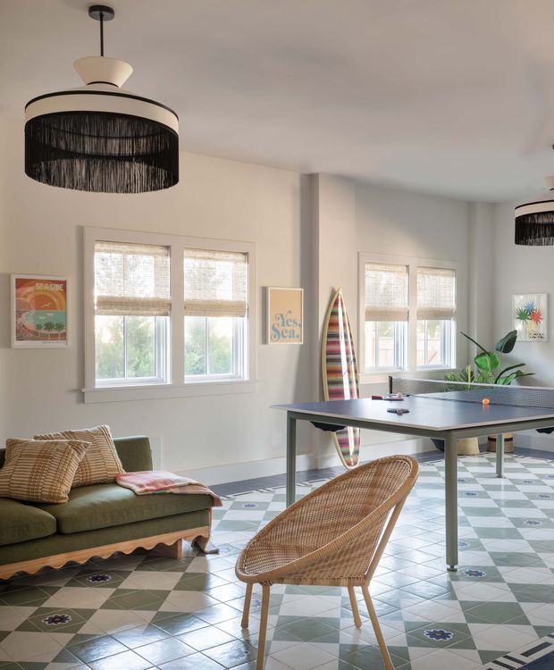 Bright, playful recreational living space combining a lounge area and table-tennis setup. The room has white walls, multiple windows with woven blinds, patterned geometric tile flooring, a green sofa, woven rattan chair, surfboard artwork and a large black woven pendant light.