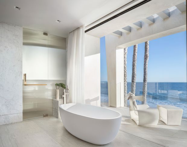 Bright, luxurious contemporary bathroom with a freestanding oval bathtub beside a frameless glass shower enclosure. The space features pale stone-like wall and floor finishes, a minimalist white palette, recessed ceiling lighting, sheer curtains, and expansive floor-to-ceiling openings overlooking the ocean. Sculptural white lounge seating and palm trunks are visible in the adjoining outdoor view.