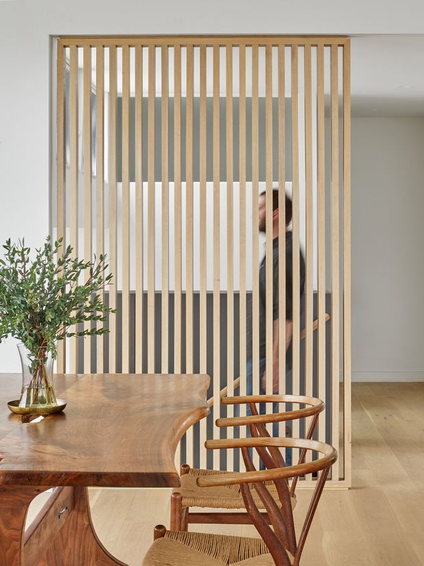 A warm contemporary dining area featuring a sculptural solid timber dining table and woven-seat timber chairs in the foreground. A full-height vertical timber slat screen forms a semi-transparent divider, with a person walking behind it. The room has pale walls, light timber flooring, and a restrained neutral palette.
