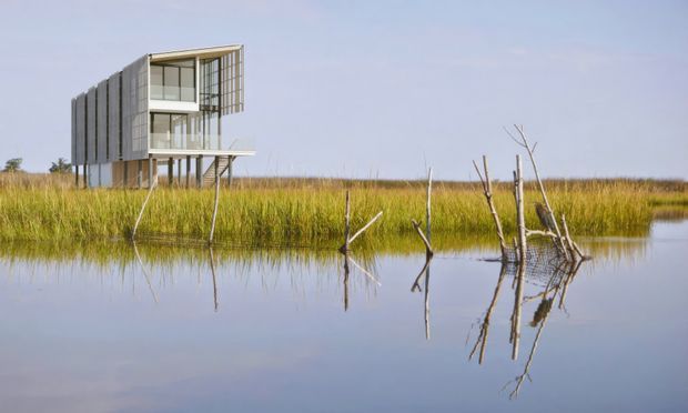 A contemporary elevated, lightweight pavilion-like building stands beside calm wetlands, with a strongly glazed upper level, projecting balcony or platform, and vertical screened facade elements. The structure is reflected in the water and surrounded by tall grasses and sparse dead branches, creating a minimalist architectural composition in a natural setting.