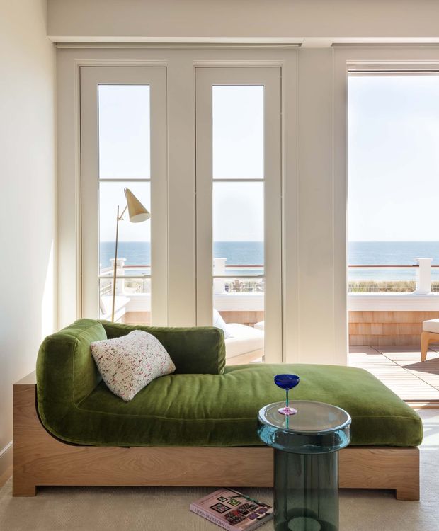 Bright coastal living room with a sculptural olive-green sofa on a timber plinth, a floral accent cushion, and a small translucent glass side table. Full-height white-framed glazed doors and windows open toward a balcony or terrace with clear ocean views. The room has pale walls, light timber flooring, and a relaxed yet refined contemporary aesthetic.