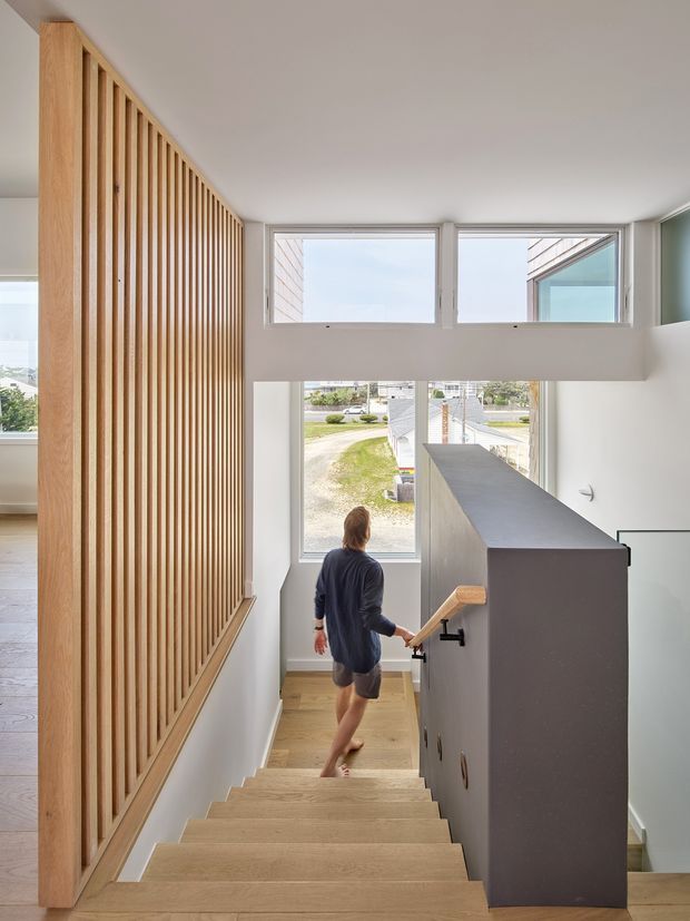 Bright contemporary stairway viewed from above, with a light timber staircase and handrail, a tall vertical timber batten screen along the left wall, and a dark grey stair balustrade or partition on the right. Large white-framed windows bring in daylight and provide views toward the street. The space has clean white walls, warm timber surfaces, and a double-height or open stairwell volume.