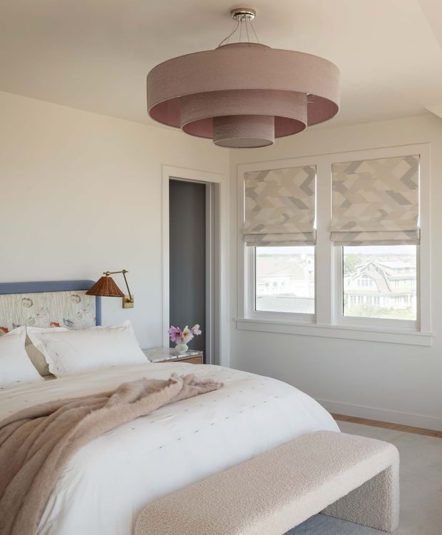 A bright, softly styled bedroom with a large upholstered bed, white bedding, a blue upholstered headboard, and a curved cream boucle bench at the foot. A sculptural layered drum pendant hangs centrally from the ceiling, while a brass wall-mounted reading lamp and small bedside table sit beside the bed. Two windows with geometric Roman blinds provide daylight and views of neighbouring homes. The room has light neutral walls, pale carpet, and a calm contemporary aesthetic.