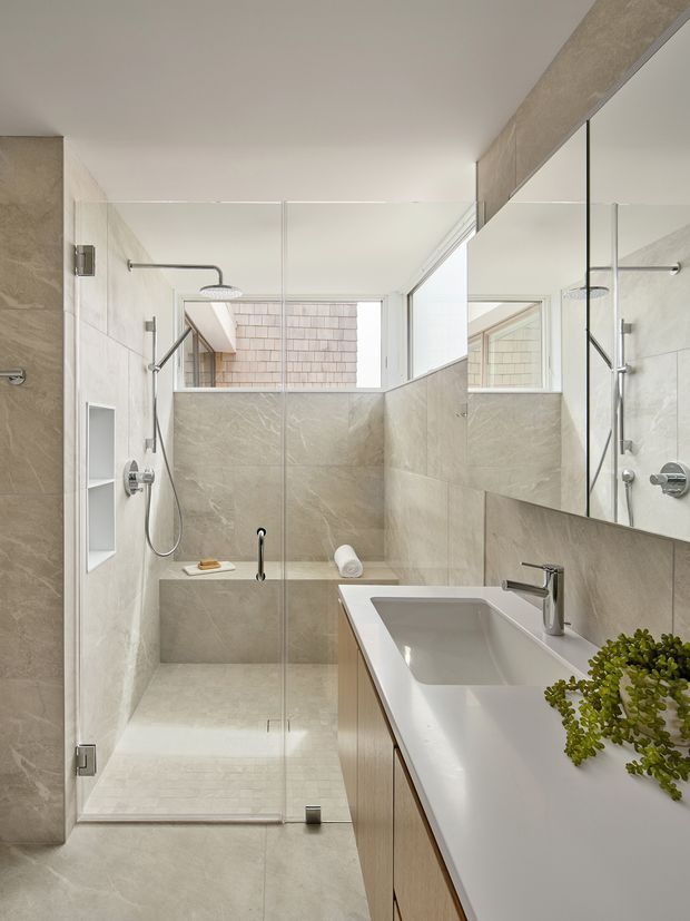 A bright, modern bathroom with a walk-in glass shower, large-format beige stone-look wall and floor tiles, a floating timber-toned vanity with an integrated white basin, chrome tapware, and a large mirror. Natural light enters through a high window, with a small plant adding greenery.