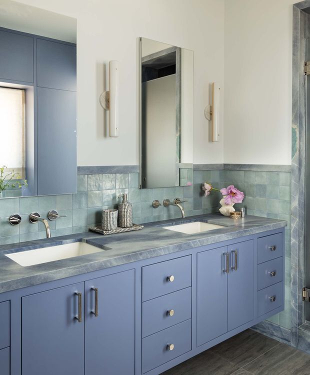 A stylish bathroom vanity with a long blue-painted built-in cabinet, dual undermount basins, brushed metal tapware, and a grey-blue stone-look benchtop. The walls feature pale blue-green square tiles with a darker horizontal trim, while a central rectangular mirror and slim vertical wall lights sit above the vanity. A glass shower enclosure is visible at right, and dark timber-look flooring grounds the room.