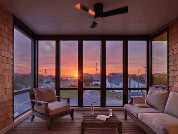 A cosy enclosed sunroom or porch with expansive black-framed glazing overlooking a suburban neighbourhood at sunset. The room features timber shingle-clad walls, a timber plank floor, a ceiling fan, and upholstered lounge seating arranged around a low wooden coffee table. Warm sunset light creates a relaxed, intimate atmosphere.
