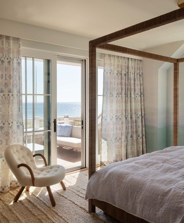 Bright coastal-style bedroom with a large upholstered bed, patterned full-height curtains, a woven natural-fibre rug, and a sculptural upholstered lounge chair. Floor-to-ceiling glazed doors open to a balcony with expansive ocean views, while a large rope-wrapped four-poster bed frame adds a distinctive textural feature.