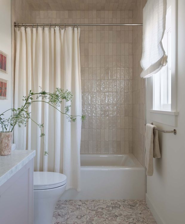 A bright, softly styled bathroom featuring a white bathtub with shower curtain, glossy cream-beige square wall tiles, a white toilet, pale vanity, patterned encaustic-style floor tiles, and a window with a relaxed Roman shade. A simple towel rail, framed artwork, and potted branch add decorative detail.