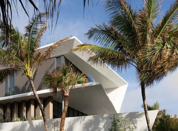Exterior architectural photograph of a striking contemporary multi-level residence partially framed by palm trees. The building has crisp white rendered volumes, dramatic angular projecting forms, deep overhangs, dark-framed glazing, and a sculptural white boundary wall. The scene has a sunny tropical or coastal character, although no specific setting is definitively identifiable.