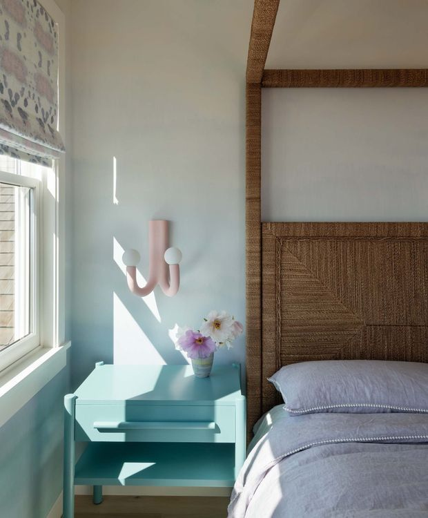 Bright, softly styled bedroom corner featuring a bed with a tall woven rattan or cane headboard, pale blue bedding, and a matching pale turquoise bedside table. A large window with a patterned Roman blind brings in strong natural light, while a sculptural blush wall light and a small vase of flowers add decorative accents. The composition has a playful, coastal-inspired feel with warm natural texture.