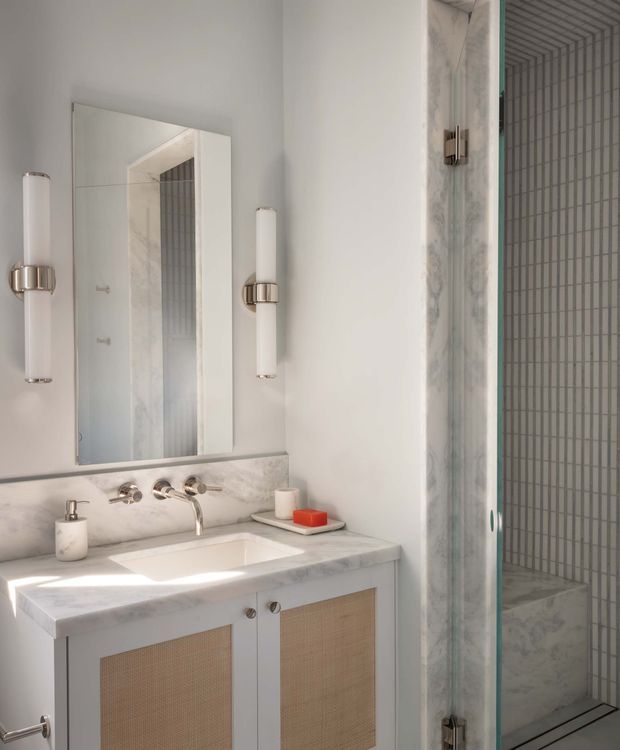 A refined contemporary bathroom vanity with a pale marble countertop and integrated rectangular basin, wall-mounted brushed metal tapware, and a mirrored wall flanked by vertical cylindrical sconces. Below, white vanity cabinetry features woven tan inset panels. At right is a frameless glass shower enclosure lined with vertically patterned grey tiles and marble trim. The room is softly lit with a light, elegant palette.