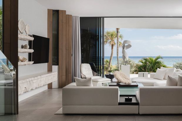 Bright contemporary living room with a refined coastal feel, furnished with low-profile cream sofas, a glass coffee table, sculptural lounge chairs, and a wall-mounted television. Full-height sliding glass doors open to a palm-fringed ocean view, while vertical timber wall panelling, a pale tiled floor, and a linear fireplace define the interior.