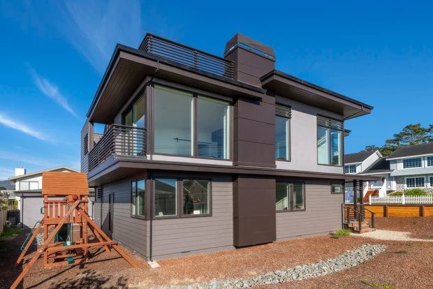 Exterior view of a contemporary two-storey residential house with dark horizontal cladding, contrasting smooth wall panels, extensive corner glazing, broad roof overhangs, and multiple metal-railed balconies. The home sits in a suburban setting with gravel landscaping and neighbouring houses visible around it.