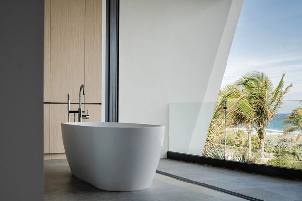 A minimalist contemporary bathroom featuring a freestanding matte white bathtub beside expansive floor-to-ceiling glazing. The room has large-format grey floor tiles, pale timber-look fixed cabinetry or wall panelling, a dark window frame, and a sculptural angled white wall reveal. Palm trees and an ocean view are visible outside.
