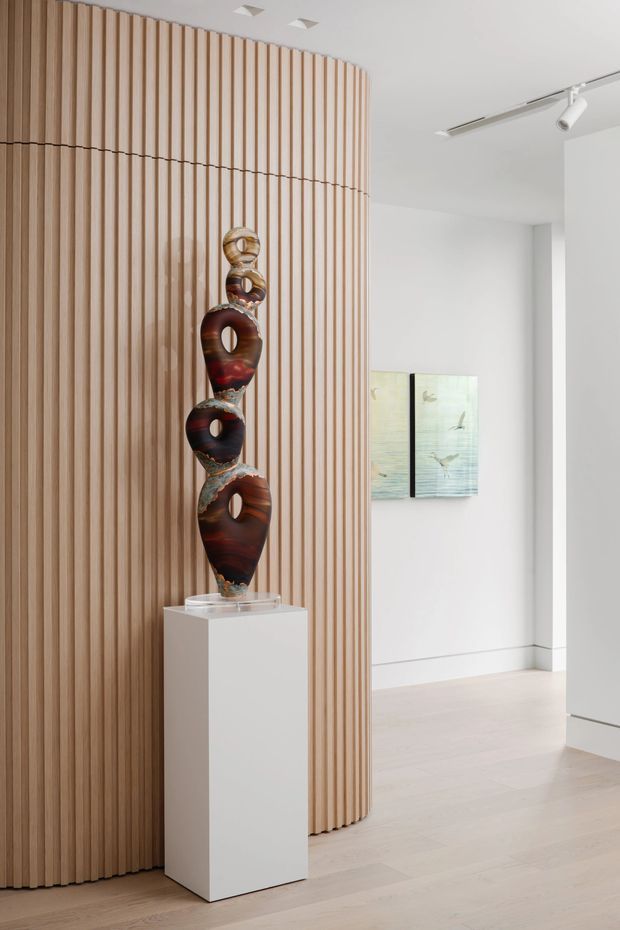 A bright, minimalist interior gallery or entry space featuring a tall sculptural artwork displayed on a white pedestal. The main architectural feature is a curved wall clad in evenly spaced vertical light timber slats, contrasting with white walls, a pale timber floor, recessed ceiling lights, and a linear track light. Artwork is also visible on the rear wall.