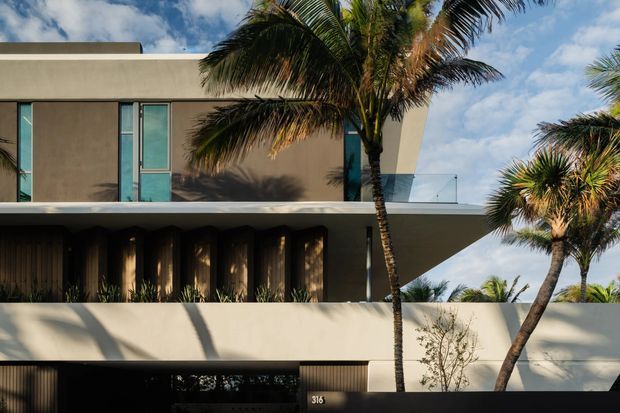 Contemporary multi-storey tropical residence with a flat roof, smooth beige rendered walls, deep overhangs, and strong horizontal massing. Narrow vertical windows and a glass-balustraded upper terrace sit above a shaded ground-level facade with vertical screening. Mature palm trees frame the building, while a dark entry wall and house number are visible at street level.