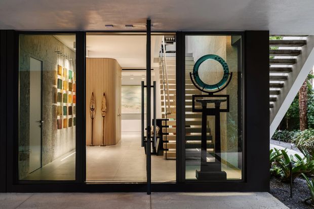 Front entrance of a contemporary residence, viewed from an exterior covered threshold. Black-framed glazed entry doors open into a refined foyer with a curved timber feature wall, floating staircase, artwork, and a prominent sculptural circular installation. A planted garden is visible beside the entry, and the flat overhanging roof creates a sheltered arrival zone.