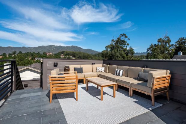A sunny rooftop terrace with a contemporary outdoor seating arrangement, including a large cushioned sectional sofa, wooden armchairs, and a low coffee table on a light outdoor rug. Dark tiled paving and charcoal privacy walls frame the terrace, with a mountain landscape and trees visible beyond the railing.