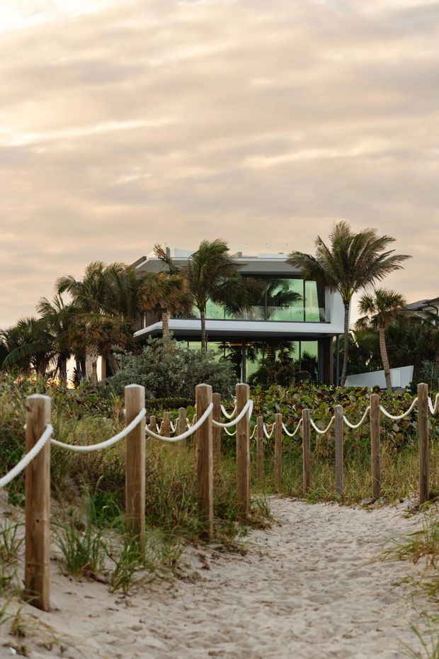 A contemporary two-storey house viewed from a sandy pathway, partially screened by lush tropical vegetation and palm trees. The building has broad floor-to-ceiling glazing, clean horizontal lines, a flat or very low-pitched roof, and a light neutral facade. Timber posts with rope define the foreground walkway, creating a relaxed coastal setting.