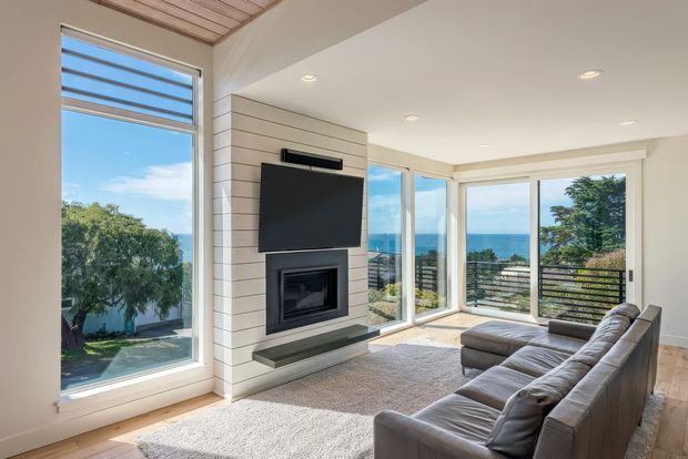 Bright contemporary living room with a large sectional sofa facing a wall-mounted television and built-in fireplace. Floor-to-ceiling corner glazing opens to expansive ocean views, while light timber flooring, a pale horizontal wall feature, recessed lighting, and a timber-lined ceiling add warmth.