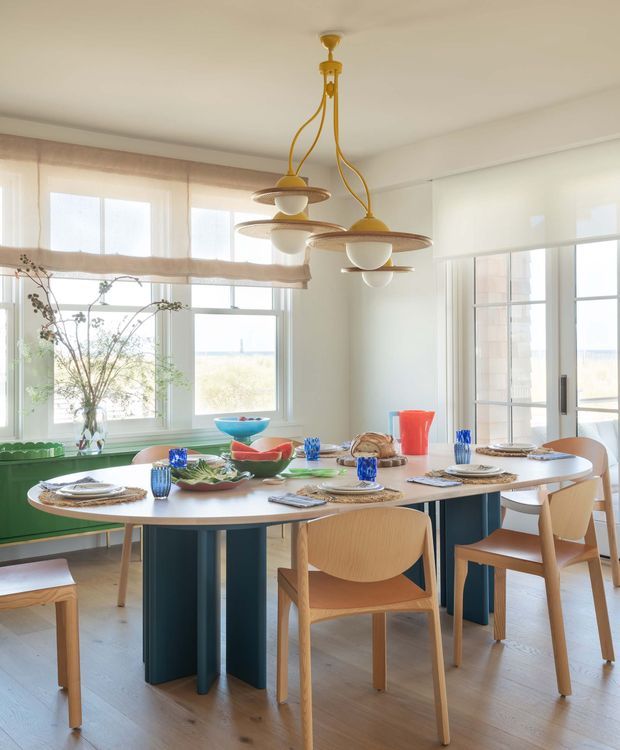Bright, airy dining room with a large oval dining table surrounded by light timber chairs. The room features pale timber flooring, white walls, expansive windows with sheer and roller blinds, and a built-in deep green bench beneath the glazing. A distinctive cluster of sculptural pendant lights hangs above the table, while blue glassware and colourful table settings provide accents. Coastal scenery is visible through the windows.