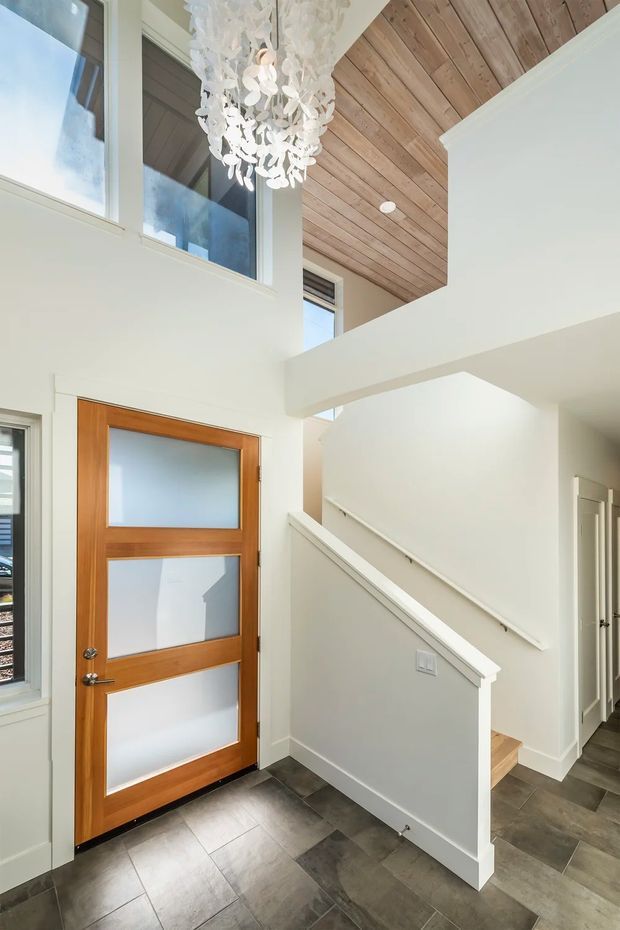 Bright contemporary residential entry hall with a timber-and-frosted-glass front door, dark large-format tiled flooring, white walls and trim, and a stairway leading upward. The space has a double-height volume with upper-level windows, a warm timber-lined ceiling section, and a prominent white sculptural pendant light.