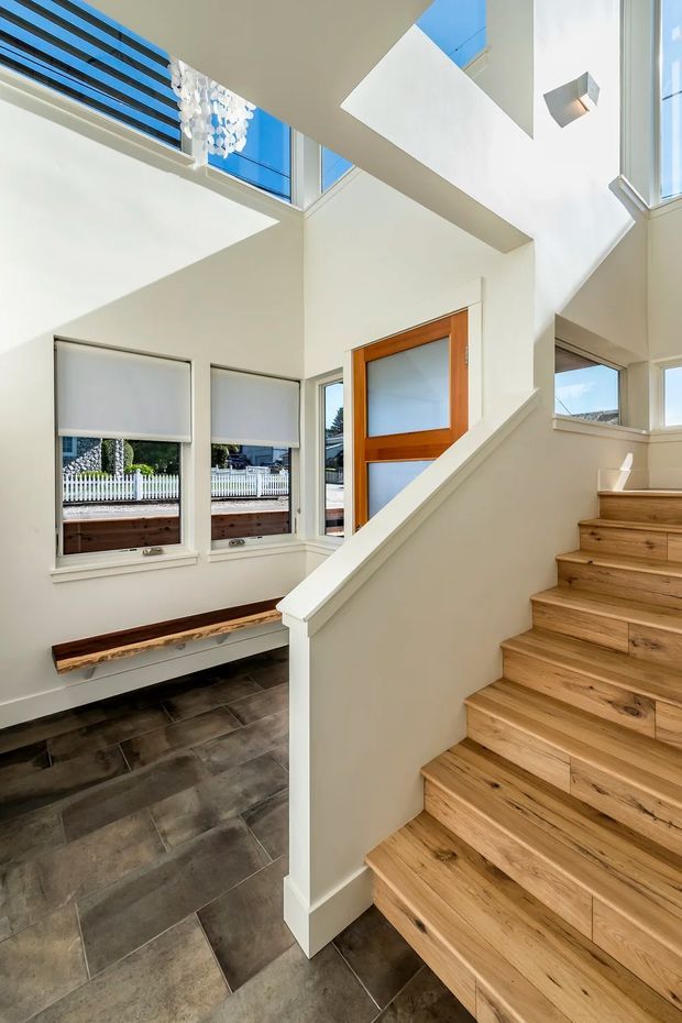 Bright contemporary residential stairway and entry hall with a timber staircase, white walls, large windows, and high clerestory glazing. The open stair volume is illuminated by strong natural light, with dark tiled flooring below and a built-in timber bench beneath the windows.