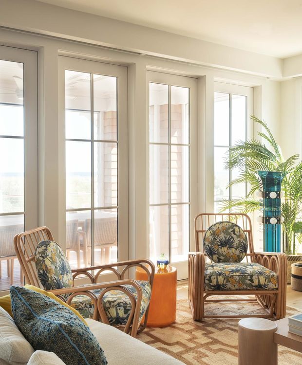 A bright, airy living room with tall multi-pane glazed doors and windows, pale walls, rattan lounge chairs upholstered in botanical floral fabric, a light sofa with patterned cushions, a geometric rug, and a tall indoor palm. Warm timber and woven textures create a relaxed, coastal-inspired seating area.