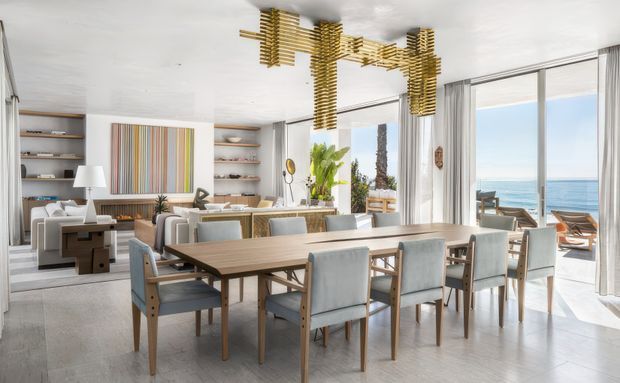 Bright open-plan dining and living area with a long timber dining table, upholstered dining chairs, built-in shelving, contemporary artwork, and a sculptural timber pendant light. Full-height glazed doors open toward a blue ocean view and bring abundant natural light into the refined coastal-feeling interior.