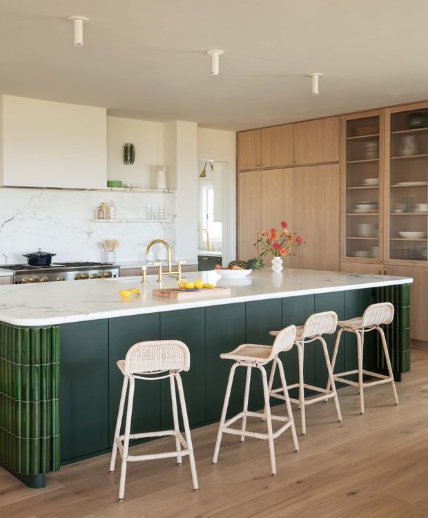 A bright, contemporary kitchen centered on a large marble-look island with a deep green fluted base and brass tapware. The island is paired with four light woven bar stools. Warm timber cabinetry, open glass-front shelving, white marble-look splashback, timber flooring, and integrated appliances create a refined yet relaxed interior. Decorative flowers and kitchenware add colour and texture.