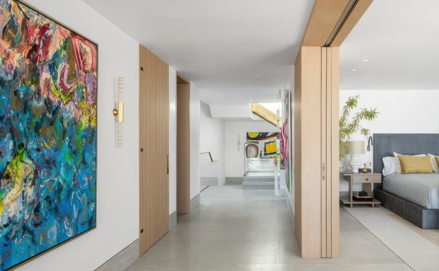A bright contemporary residential hallway with pale timber doors and wall panelling, light grey flooring, white walls and ceiling, and large colourful abstract artwork. A bedroom is visible through an open doorway on the right, while reflective glazing or a mirror extends the view toward additional artwork and circulation space.