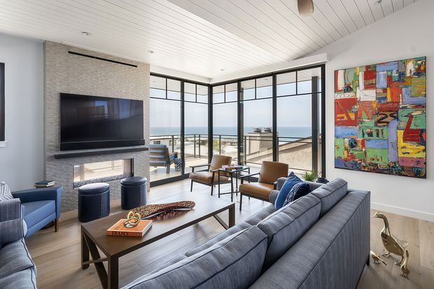 Bright contemporary living room with a large sectional sofa, lounge chairs, coffee table, fireplace and wall-mounted television. Full-height black-framed glazing opens the room visually to an ocean-facing terrace, while a colourful abstract artwork provides a strong accent. The room has a white linear-paneled ceiling and neutral textured finishes.