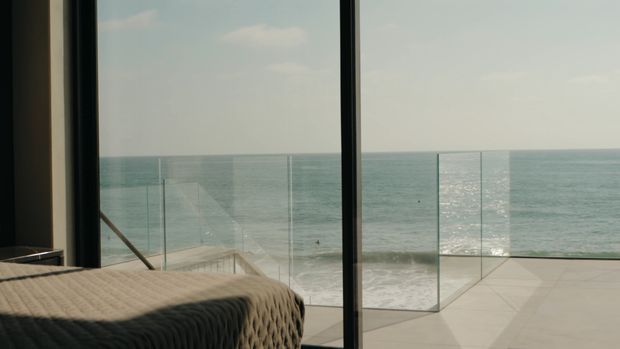 A bedroom interior opens directly onto a glass-railed terrace overlooking the ocean. The foreground shows the corner of a textured bed, while full-height dark-framed glazing provides expansive coastal views. The terrace has pale large-format paving and frameless glass balustrades, creating a minimalist, airy composition.