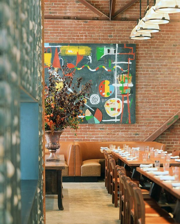 A warmly lit restaurant dining area with exposed red-brick walls, dark structural steel bracing, a polished concrete floor, and a row of suspended industrial pendant lights. A long communal dining table is set with glassware and white napkins, alongside a caramel leather banquette and wooden chairs. A large colourful abstract artwork and a tall floral arrangement add bold decorative accents.