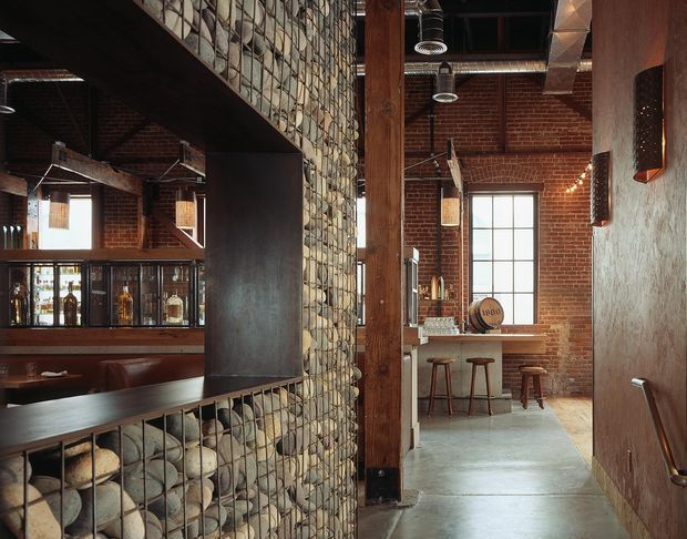 An industrial-style bar and hospitality interior with exposed red brick walls, dark timber framing, polished concrete flooring, and a distinctive gabion-style partition filled with natural stones. A long dark bar counter with leather seating and bottle shelving occupies the left side, while a compact high table and stools sit near a tall multi-pane window at the rear. Exposed ductwork, pendant lighting, timber posts, and metal details reinforce the rugged warehouse character.